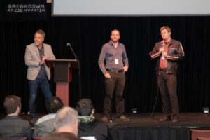 Three people stand on a stage, with a projector screen behind them, looking out into the audience.